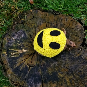 Könnte beinhalten: Eine kleine Schildkröte trägt ein gelbes, gehäkeltes Smiley-Kostüm. Die Schildkröte sitzt auf einem Baumstumpf.