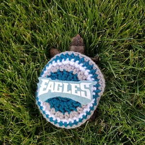 以下が含まれることがあります： 白い文字で「EAGLES」と書かれた緑色のフェルトパッチと、白とグレーのストライプの入ったかぎ針編みの帽子をかぶった小さなカメ