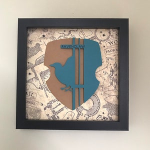May include: A framed print of the Ravenclaw house crest from the Harry Potter series. The crest features a blue bird with a brown background and the text "Ravenclaw" in white. The frame is black and the background is a map of Hogwarts.