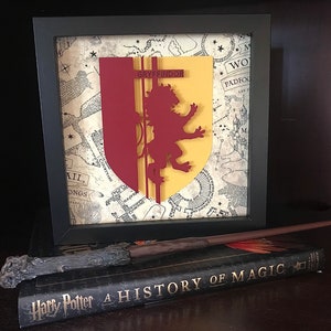 May include: A framed Gryffindor house crest from the Harry Potter series, featuring a red lion on a yellow shield, against a background of a parchment map. The frame is black and the crest is 3D.