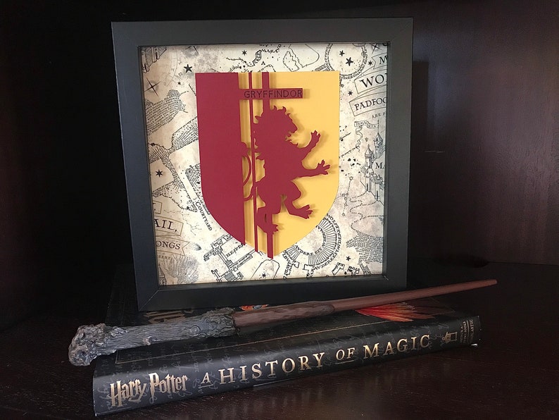 May include: A framed Gryffindor house crest from the Harry Potter series, featuring a red lion on a yellow shield, against a background of a parchment map. The frame is black and the crest is 3D.