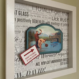 May include: A framed collage with a vintage-style sign that reads "Welcome to Schitt's Creek" and a photo of a couple in a snowy setting. The collage is surrounded by a white frame and a background of text that includes the words "TOTALLY get that", "LICK RUST", and "BEDEVILED". A red arrow points to a note that reads "Don't worry. It's his sister!"