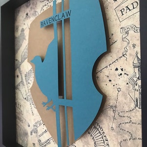 May include: A framed 3D papercraft of the Ravenclaw house crest from the Harry Potter series. The crest is blue and brown with a bird and the words "Ravenclaw" on it. The background is a map of Hogwarts.