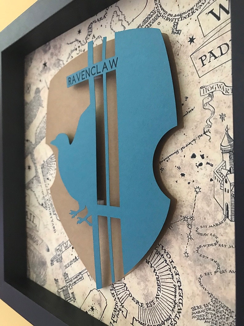May include: A framed 3D papercraft of the Ravenclaw house crest from the Harry Potter series. The crest is blue and brown with a bird and the words "Ravenclaw" on it. The background is a map of Hogwarts.