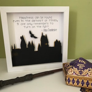 May include: A white framed picture with a black silhouette of Hogwarts castle and a quote from Albus Dumbledore: "Happiness can be found even in the darkest of times. If one only remembers to turn on the light."  A wand and a box of chocolate frogs are in the foreground.