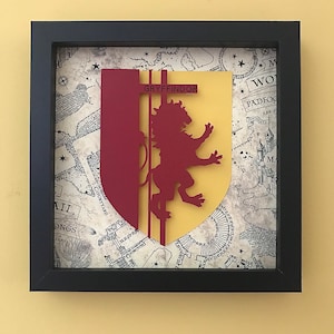 May include: A framed papercraft of the Gryffindor house crest from the Harry Potter series. The crest features a red lion on a yellow background with the word "Gryffindor" in black text. The background is a map of Hogwarts.