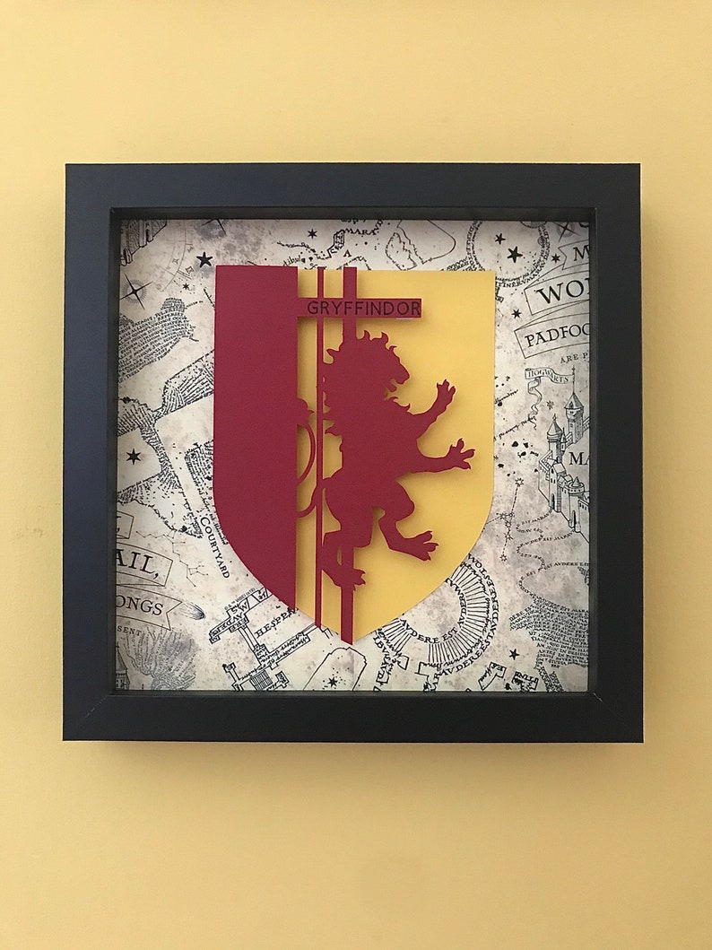 May include: A framed papercraft of the Gryffindor house crest from the Harry Potter series. The crest features a red lion on a yellow background with the word "Gryffindor" in black text. The background is a map of Hogwarts.