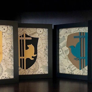 May include: Four framed prints of the Hogwarts house crests. Each crest is a different color and features a different animal: a lion, a badger, a snake, and an eagle. The crests are on a background of a parchment-like map.