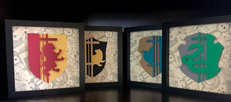 May include: Four framed prints of the Hogwarts house crests. Each crest is a different color and features a different animal: a lion, a badger, a snake, and an eagle. The crests are on a background of a parchment-like map.
