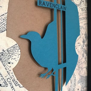 May include: A brown paper cutout of the Ravenclaw house crest from the Harry Potter series. The crest features a blue raven with a silver beak and legs, perched on a silver branch. The words "RAVENCLAW" are printed in black on a silver banner above the raven.