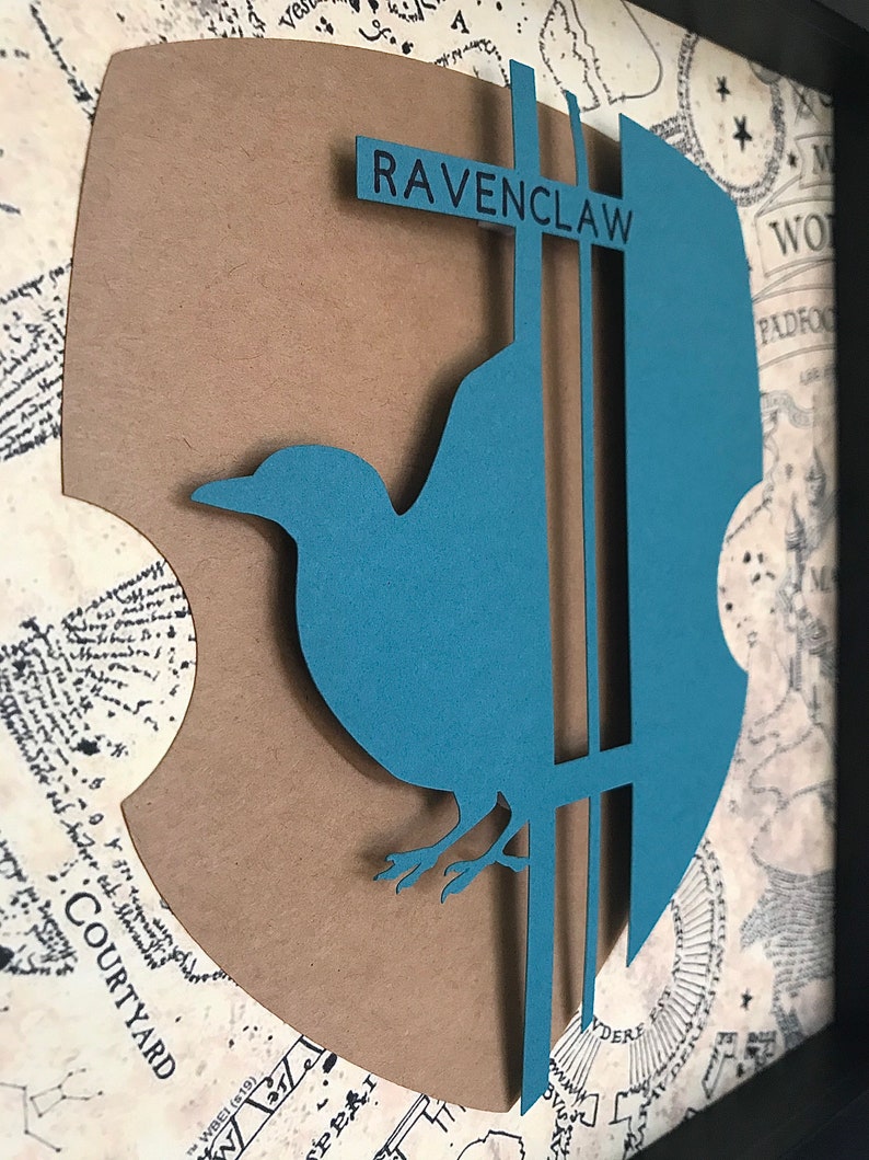 May include: A brown paper cutout of the Ravenclaw house crest from the Harry Potter series. The crest features a blue raven with a silver beak and legs, perched on a silver branch. The words "RAVENCLAW" are printed in black on a silver banner above the raven.