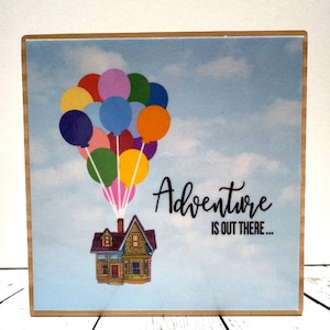 May include: A wooden block with a printed image of a house flying with colorful balloons in a blue sky with white clouds. The text "Adventure is out there..." is printed below the house.