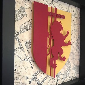 May include: A framed papercraft of the Gryffindor house crest from the Harry Potter series. The crest features a red lion on a yellow background with the word "GRYFFINDOR" in black lettering.