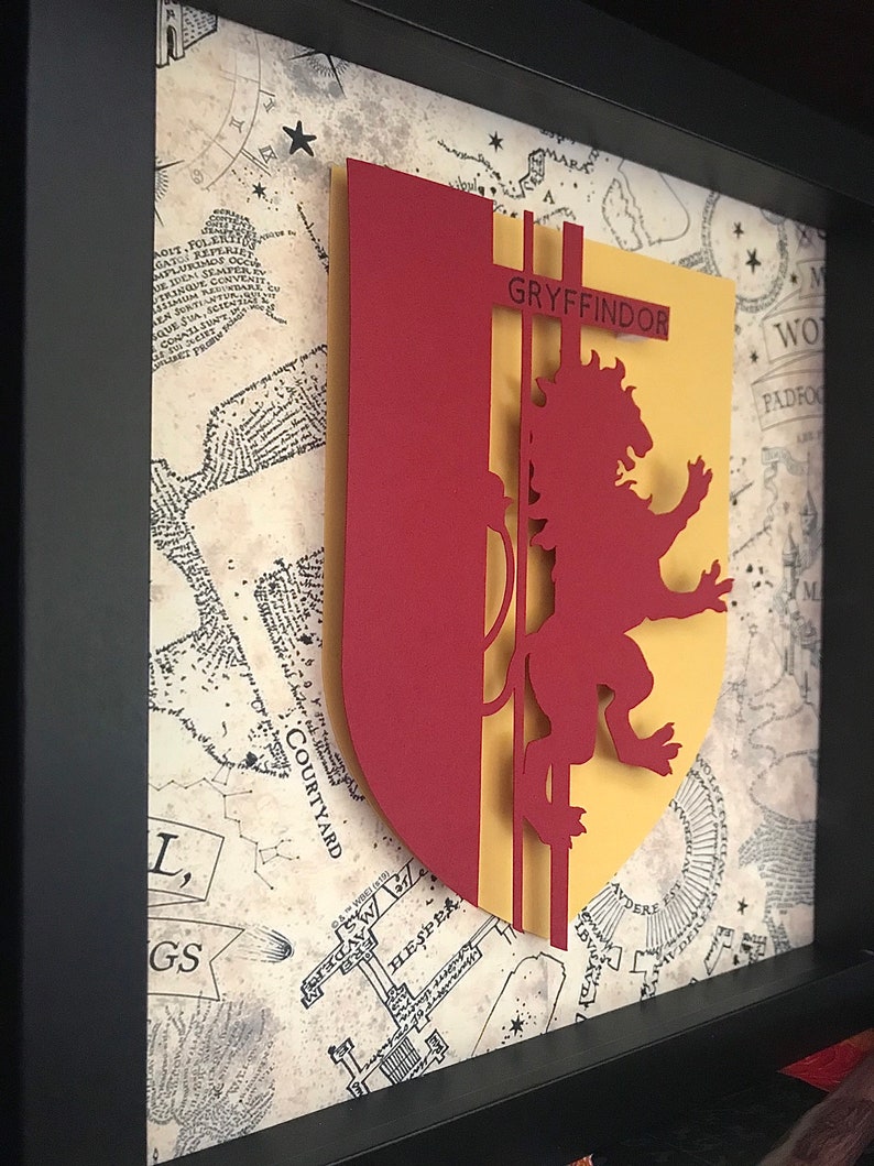 May include: A framed papercraft of the Gryffindor house crest from the Harry Potter series. The crest features a red lion on a yellow background with the word "GRYFFINDOR" in black lettering.