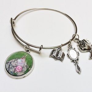May include: A silver bangle bracelet with four charms: a green round charm with a picture of a castle, a silver book charm, a silver mirror charm, and a silver rose charm.
