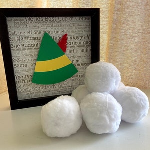 May include: A framed print with text, an elf hat, and several white, fluffy snowballs. The green elf hat has a yellow band and a red feather. The framed print has a black frame and a background of text.