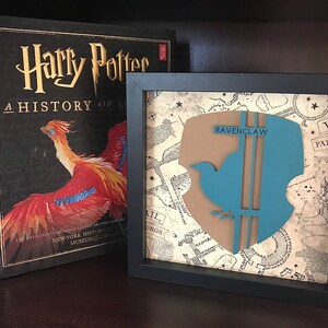 May include: A framed print of the Ravenclaw house crest from the Harry Potter series. The crest features a blue bird with a brown background and the words "Ravenclaw" in white text. The crest is displayed on a background of a vintage map.