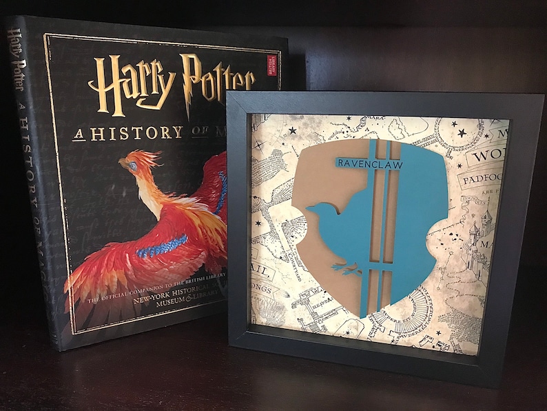 May include: A framed print of the Ravenclaw house crest from the Harry Potter series. The crest features a blue bird with a brown background and the words "Ravenclaw" in white text. The crest is displayed on a background of a vintage map.