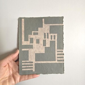 Puede incluir: Una obra de arte en papel hecha a mano con un dise&ntilde;o geom&eacute;trico. La obra tiene un fondo gris con un dise&ntilde;o de color beige claro recortado. Los bordes del papel son &aacute;speros e irregulares, lo que se suma a la est&eacute;tica artesanal. La obra es sostenida por una mano.