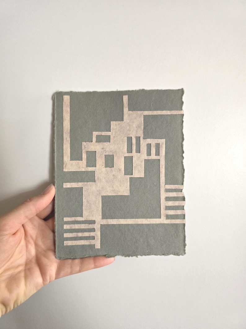 Puede incluir: Una obra de arte en papel hecha a mano con un dise&ntilde;o geom&eacute;trico. La obra tiene un fondo gris con un dise&ntilde;o de color beige claro recortado. Los bordes del papel son &aacute;speros e irregulares, lo que se suma a la est&eacute;tica artesanal. La obra es sostenida por una mano.