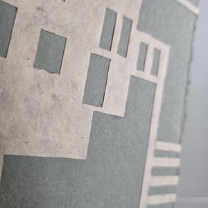 Puede incluir: Primer plano de una obra de arte en papel hecha a mano. La obra presenta un dise&ntilde;o geom&eacute;trico con formas recortadas en color beige claro, superpuestas sobre un fondo verde azulado apagado. Los bordes del papel son &aacute;speros, lo que le da un aspecto texturizado.