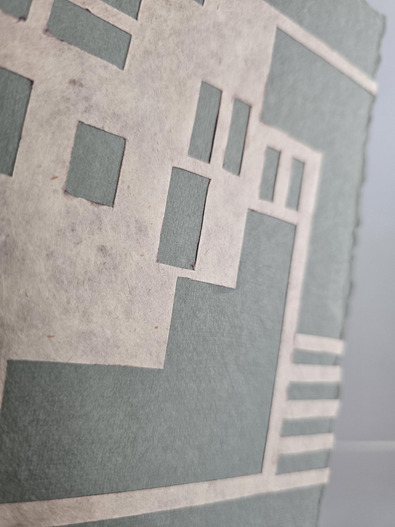 Puede incluir: Primer plano de una obra de arte en papel hecha a mano. La obra presenta un dise&ntilde;o geom&eacute;trico con formas recortadas en color beige claro, superpuestas sobre un fondo verde azulado apagado. Los bordes del papel son &aacute;speros, lo que le da un aspecto texturizado.