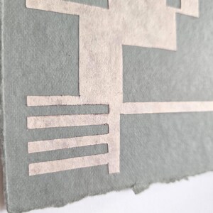 Puede incluir: Primer plano de una obra de arte en papel hecha a mano. La obra presenta un dise&ntilde;o geom&eacute;trico recortado en papel de color claro, sobre un fondo verde apagado. Los bordes del papel son &aacute;speros y desiguales.