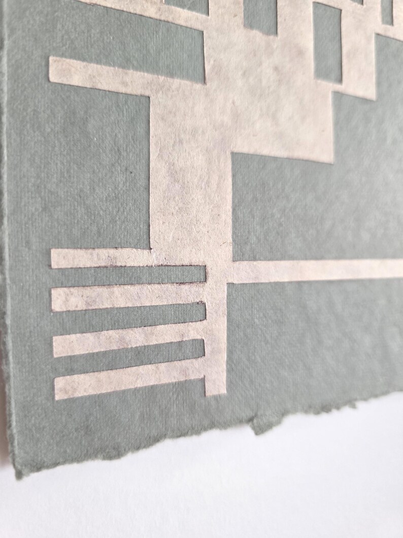 Puede incluir: Primer plano de una obra de arte en papel hecha a mano. La obra presenta un dise&ntilde;o geom&eacute;trico recortado en papel de color claro, sobre un fondo verde apagado. Los bordes del papel son &aacute;speros y desiguales.