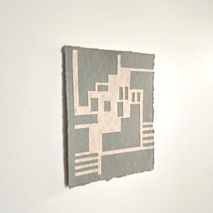 Puede incluir: Una obra de arte rectangular gris con un dise&ntilde;o geom&eacute;trico en rosa claro. La obra de arte tiene una superficie rugosa y texturizada y un borde rasgado. El dise&ntilde;o presenta l&iacute;neas rectas y formas rectangulares.