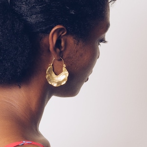 May include: A woman with dark curly hair wearing a gold hoop earring. The earring is a large, textured, crescent shape.