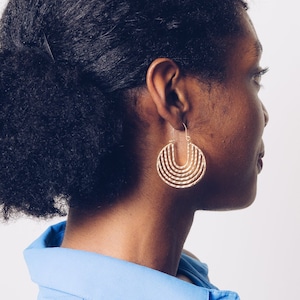 May include: A person with dark curly hair wearing a blue shirt and gold hoop earrings. The earrings are made of multiple thin bands of metal that form a semi-circle shape.