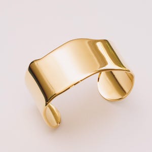 May include: A gold-colored cuff bracelet with a smooth, reflective surface. The bracelet has a curved, open design, with rounded edges and a polished finish. The jewelry piece is set against a plain background, highlighting its metallic sheen.