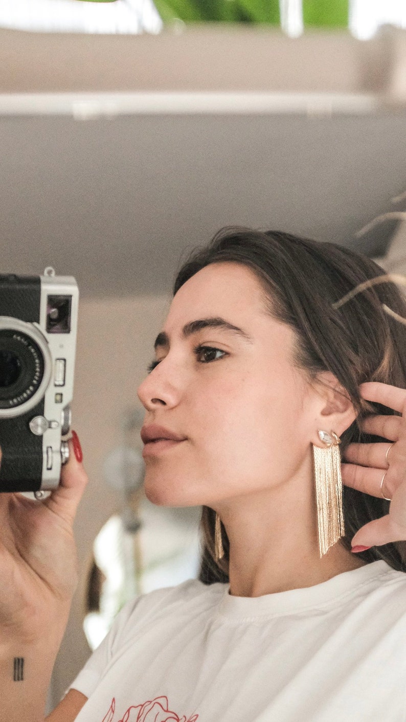 gold chain long chain fringe earrings with crystal marquis stud on model with brown hair holding a vintage camera and taking picture of reflection in the mirror