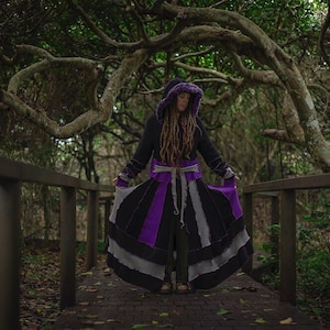 May include: A long black coat with a purple and grey skirt is worn on a wooden bridge in a forest. The coat has a hood with purple trim and a matching belt. The skirt has vertical stripes of black, purple, and grey.