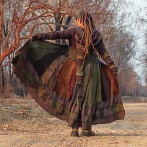 May include: A long patchwork coat in shades of brown, green, and rust is displayed outdoors. The coat has a hooded design and is worn with matching trousers. The person is standing on a dirt path with trees in the background.
