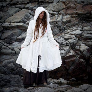 May include: A long, white hooded coat with a flowing skirt, featuring various textured knit panels. The coat has long sleeves and a button closure. The person is wearing a long brown skirt and standing in front of a rocky background.