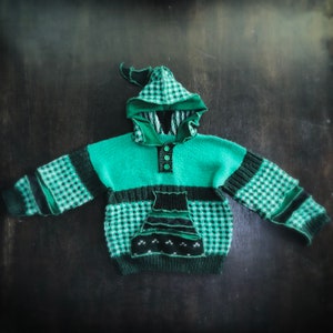 May include: A green and black knitted hoodie with a unique design. The hoodie features a checkered pattern on the sleeves and hood, a dark green pocket, and three buttons. The hood has a pointed shape.