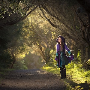 May include: A colourful hooded coat in purple, teal, and green is worn on a forest path. The coat has a unique layered design and long sleeves. The person is wearing black boots. The path is surrounded by trees.