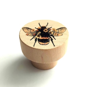 May include: A round, light-coloured wooden knob featuring a printed graphic of a bumblebee adorned with small yellow flowers on its back. The bee has black and yellow stripes, black legs, and translucent wings, centred on the flat top surface.