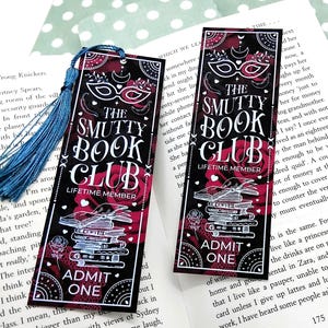 Könnte beinhalten: Zwei schwarze Lesezeichen mit dem Text "THE SMUTTY BOOK CLUB" in Weiß. Die Lesezeichen haben ein rotes Blumendesign, eine Maske und die Worte "ADMIT ONE". Eine blaue Quaste hängt von der Oberseite eines Lesezeichens.