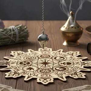 May include: A wooden divination board with engraved symbols and text, with a silver pendulum hanging above it. A brass incense burner emits smoke, and a bundle of sage is visible. The scene is set on a wooden surface.