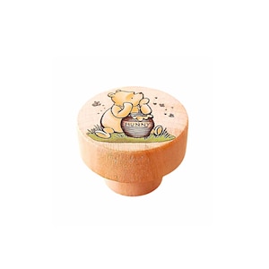 May include: A round, light-coloured wooden furniture knob featuring a printed illustration of a yellow bear sitting on green grass next to a brown honey pot labelled HUNNY. The design includes small bees flying around the bear on the flat top surface.