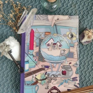 Op de afbeelding: Een notitieboek met een kleurrijke illustratie van een wereldbol, huizen en mystieke objecten. De kaft heeft een paarse rug. De afbeelding bevat ook kristallen, een glazen pot, sleutels en wierookstokjes, wat een magisch thema creëert.