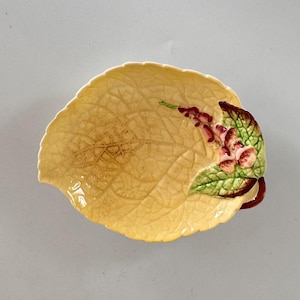 Peut inclure: Un plat en céramique en forme de feuille, avec une base jaune et des motifs de veines détaillés. Le plat présente une grappe de fleurs roses et rouges avec des feuilles vertes. Une petite poignée marron est visible.