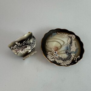 May include: A decorative teacup and saucer set. The cup and saucer are black with a white dragon design. The saucer has a gold rim and a scalloped edge. The dragon has blue accents.