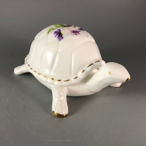 May include: A white porcelain turtle trinket box with a gold trim and a painted design of purple flowers.