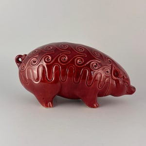 May include: A glossy, red ceramic pig figurine with a decorative swirl pattern across its back. The pig has a small curled tail and is posed with its snout pointed forward. The figurine is a decorative item.
