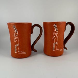 May include: Two terracotta-colored ceramic mugs with brown handles. Each mug features a white illustration of a kneeling figure with a headdress. The mugs have a textured surface and a curved handle.