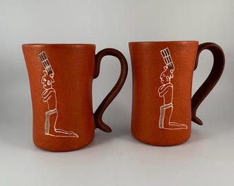 Puede incluir: Dos tazas de cerámica color terracota con asas marrones. Cada taza presenta una ilustración blanca de una figura arrodillada con un tocado. Las tazas tienen una superficie texturizada y un asa curvada.