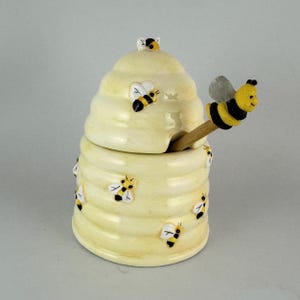 May include: A ceramic honey pot shaped like a beehive, with a lid and a wooden honey dipper. The pot is light yellow with black and yellow bee decorations. The dipper has a bee figurine on the end.
